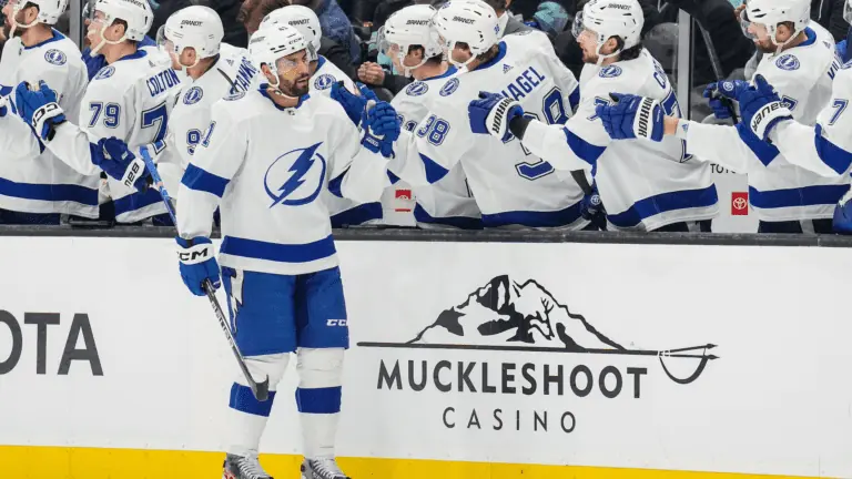 Hockey players stand up on the bench. One player is on the ice and high-fiving his teammates.