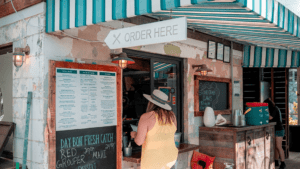 A walk up ordering window. A menu is hand written on a chalkboard to the left of the ordering window. A woman wearing a fedora is ordering.