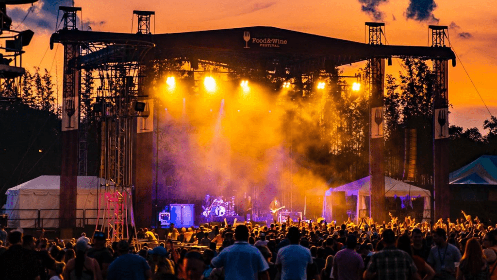 A large stage set up at sunset at a big theme park. A large crowd forms around a band.