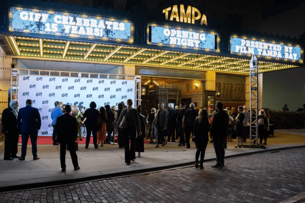 Exterior of a theatre at night. People gather under the marquee before a show.