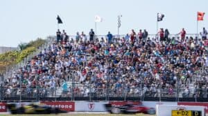 fans in bleachers watch as cars race by on a sunny day. It's an INDYCAR circuit track