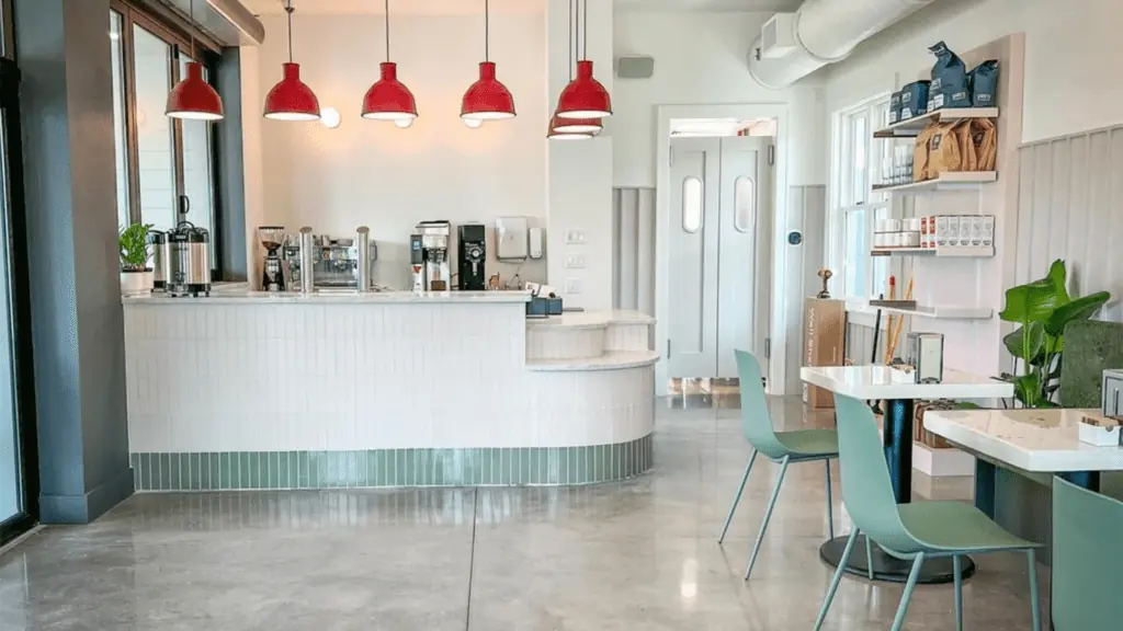 interior of a new bagel and pastry shop. There's an l shaped counter with white tiles, and hanging overhead lights with red covers. small tables with plants are arranged on the right.