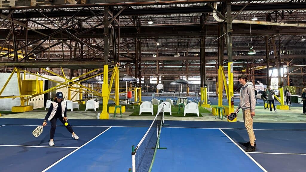 Two people volley a yellow pickleball on a court inside a warehouse