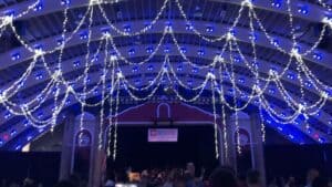 array of string lights hang over a large stage with a banner waving above. People gather around the stage. Blue lights are lit up in the rafters.