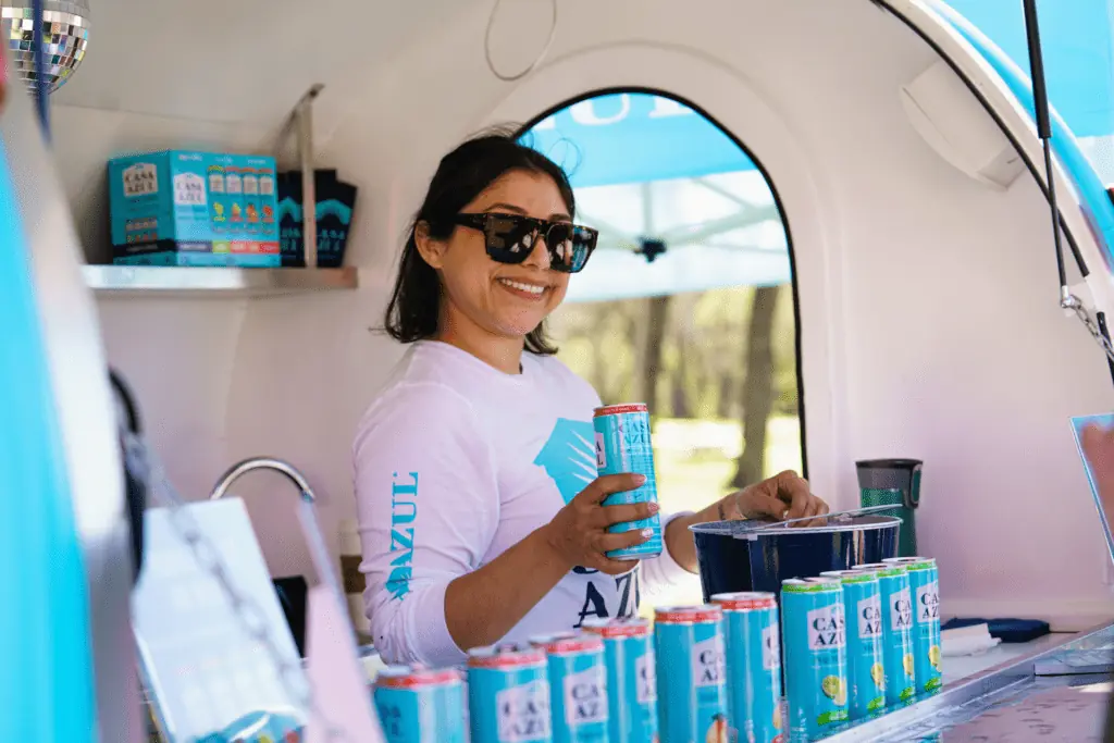 A person inside of a small car holds a blue can of tequila soda. She's wearing sunglasses and a long sleeves white shirt. Several cans of soda are lined up in the front of the cart. 