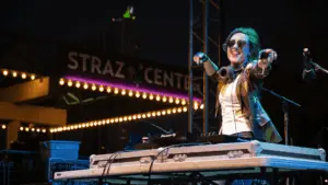 A dj on the stage at night. They're pointing to the left of the stage. A marquee is visible in the background.