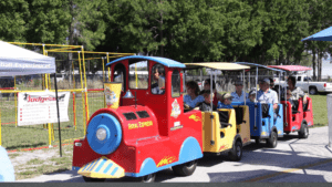 A little train chugs along a parking area. The cars are red and yellow.