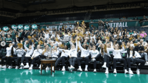 A group of fans on the basketball court celebrating. Attendees are wearing white long sleeve shirts with USF Bulls written on them. The court includes a green baseline.