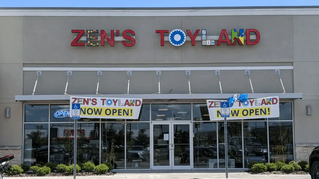 Exterior of a toy shop with a large red letter sign over the automatic doors on the ground level. Now open banners hang over the side window of the store.