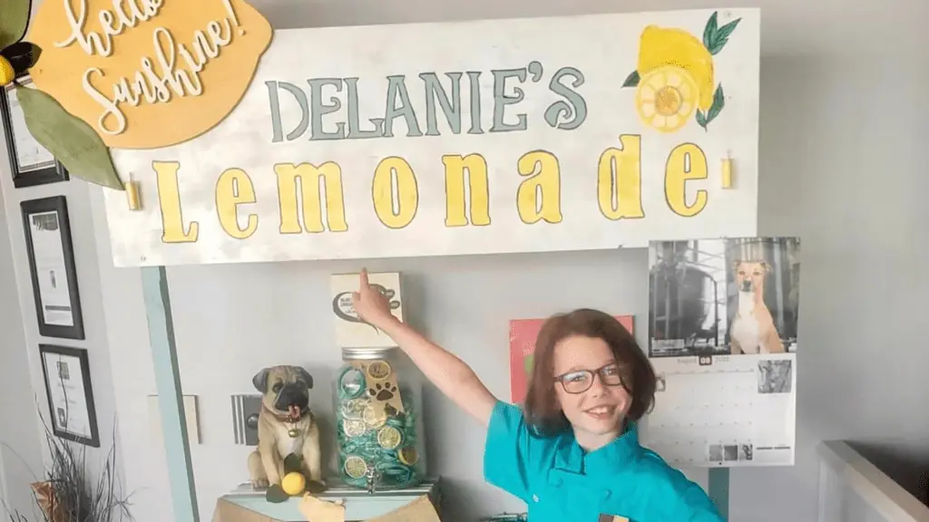 a young girl stands in front of a wooden lemonade stand sign.