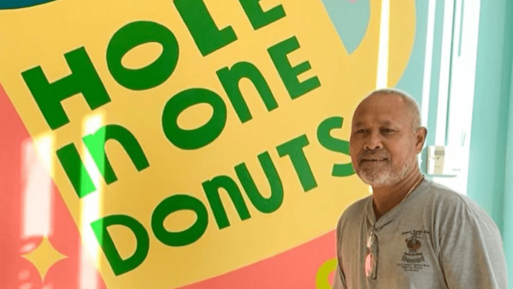 A man stands in front of a colorful mural inside of a donut shop.