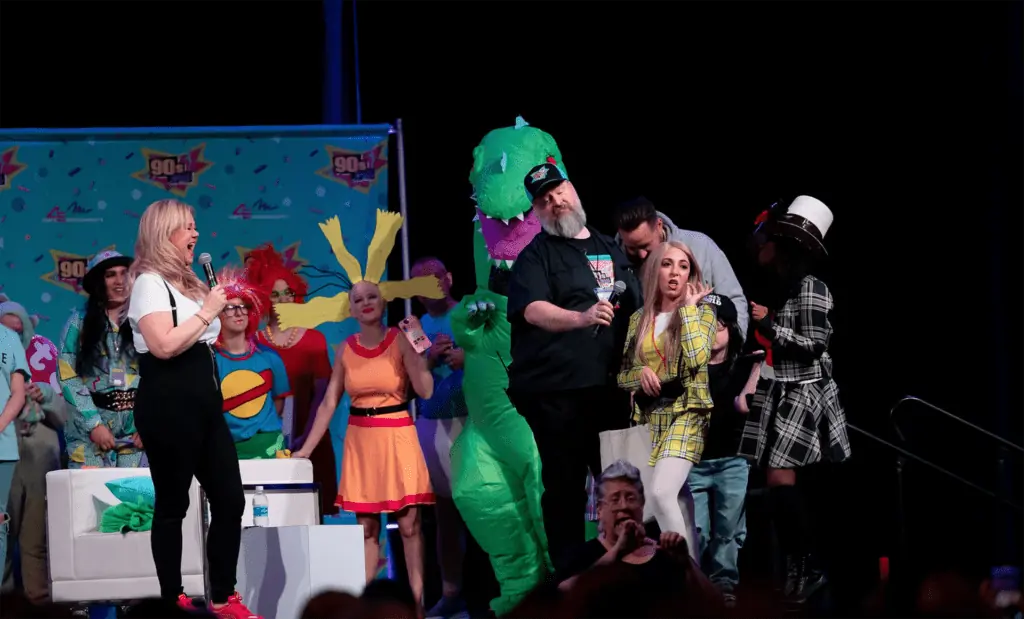 guests in costumes are gathered on a small stage for a contest. One person is dress as a giant green dinosaur