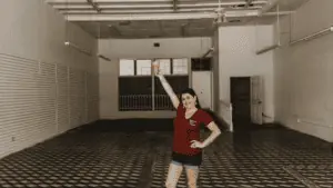 a women poses in the center of a vacant building. The woman is wearing a dark red shirt. The floor tiles have a distinct marbled pattern.