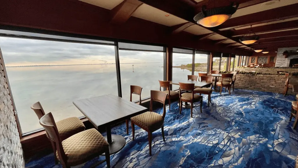 a waterfront dining room with blue carpet and tables set in front of a floor to ceiling window.