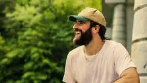 Sam Hunt wearing a white shirt and hat smiles while outside