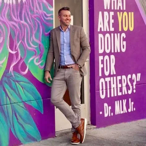 White man in brown suit standing in front of purple mural with a Dr. MLK Jr auto "What are you doing for others?"