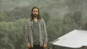 Noah Kahan in a t-shirt and cardigan standing in front of smokey trees and hills