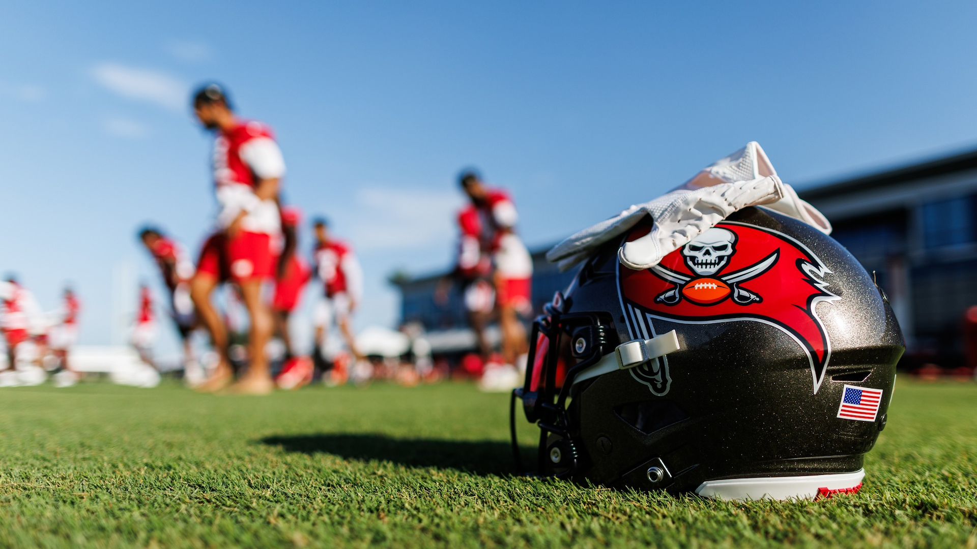 A buccaneers helmet placed on the field with a white glove over it. Football players are practicing in the background.