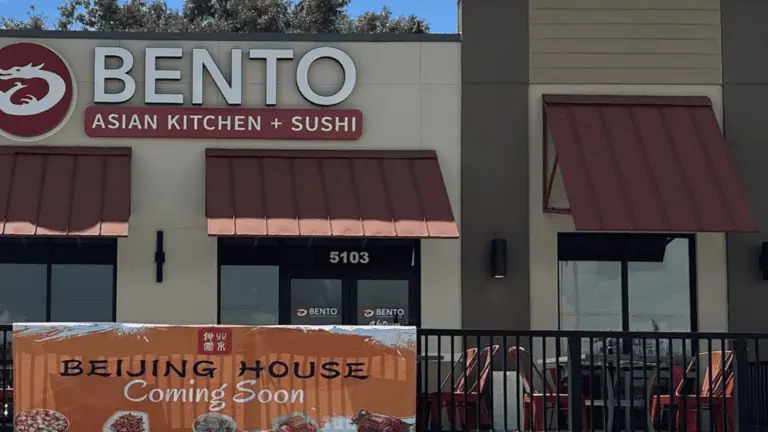 Exterior of a strip plaza restaurant with outdoor seating. A large BENTO sign is over the front door. An orange banner with a coming soon sigh is draped over the railing.