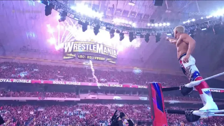 a man standing on a turnbuckle pointing at a sign that reads Wrestlemania as fireworks go off.