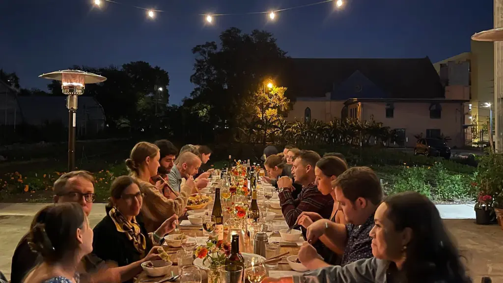 a group of people at a long dining table outside at night surrounded by a lush garden