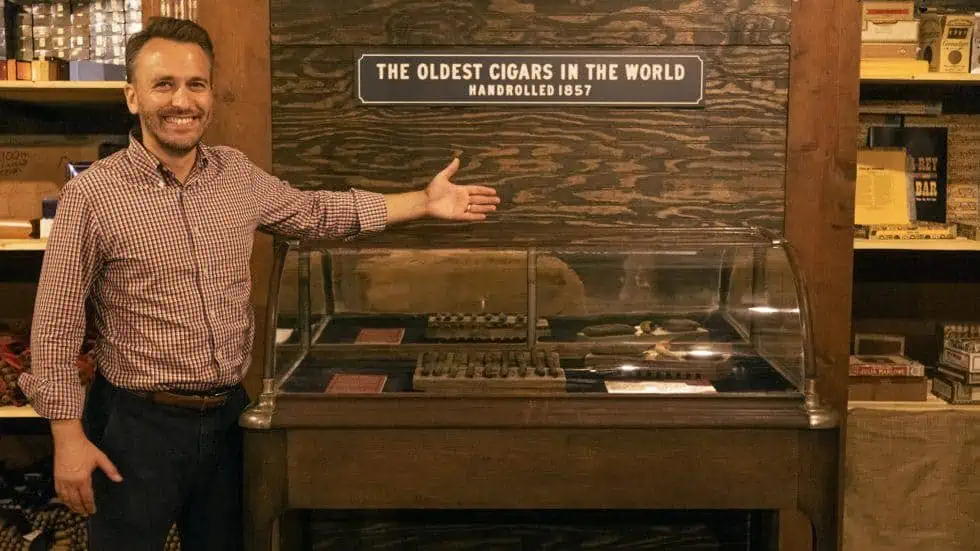 A cigar purveyor stands in front of a glass case containing the oldes cigars in the world. The setting is rustic, an aged “cigar vault” beneath a full factory.