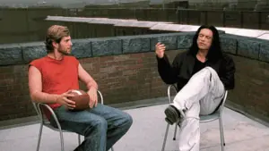 two men sitting on a rooftop talking. One is wearing a red tank top and holding a football. The other has long black hair and is wearing khaki pants and a black button down shirt.