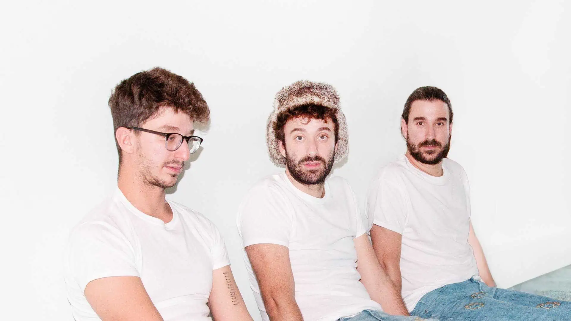 The three brothers of AJR wearing white tshirts leaning against a white wall while seated on the floor