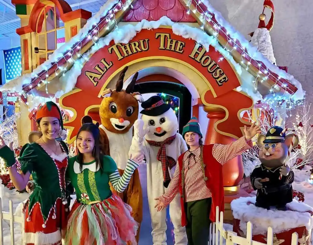 multiple holiday characters stand in front of a large gingerbread house