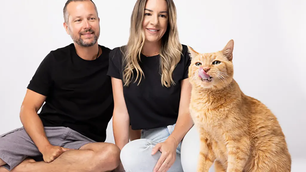 two people sit next to each other. an orange cat licks its lips in front of them