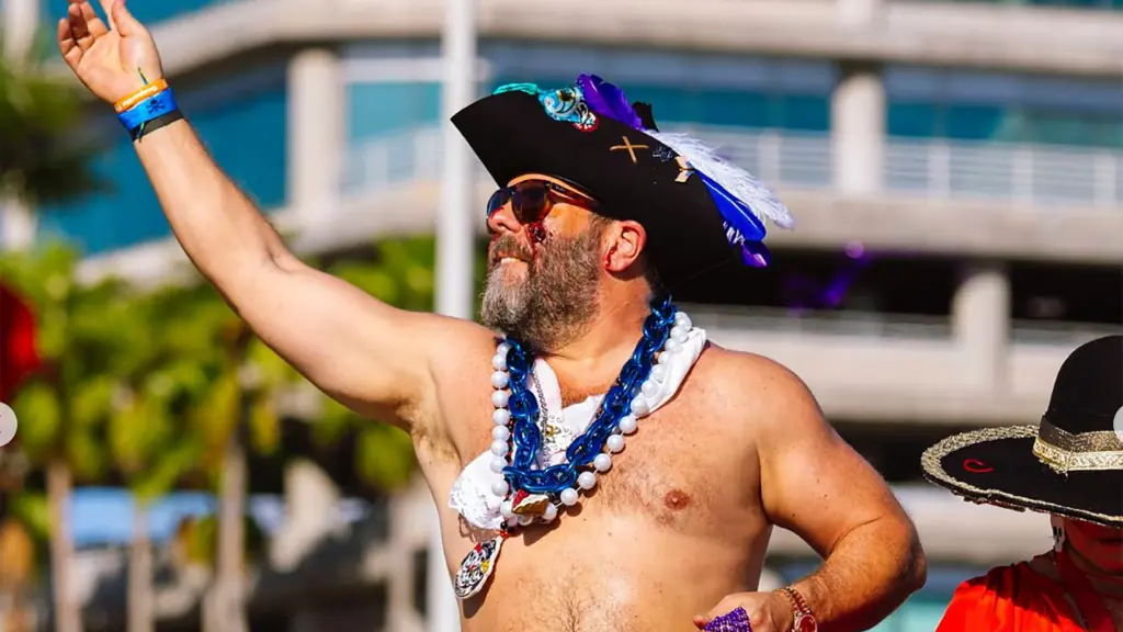 comedian in a pirate hat wears blue beads around his neck, a pirate hat, and sunglasses while tossing beads to onlookers during a parade