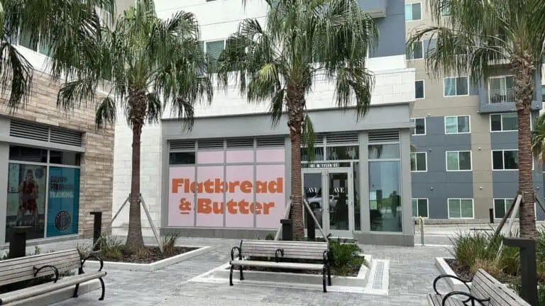 exterior of a tall building with a pink and orange sign in the window. There's a courtyard filled with palm trees and wooden benches