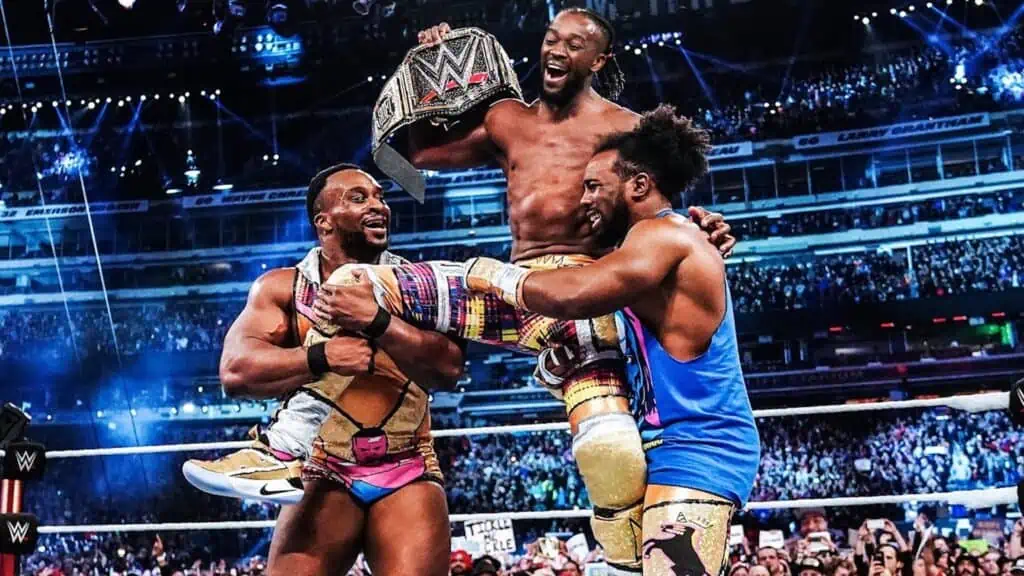 three wrestling performers pose with a big belt in the center of a ring