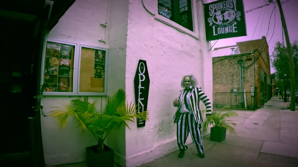 a man in a black striped suit and a green wig poses in front of a bar in a downtown area