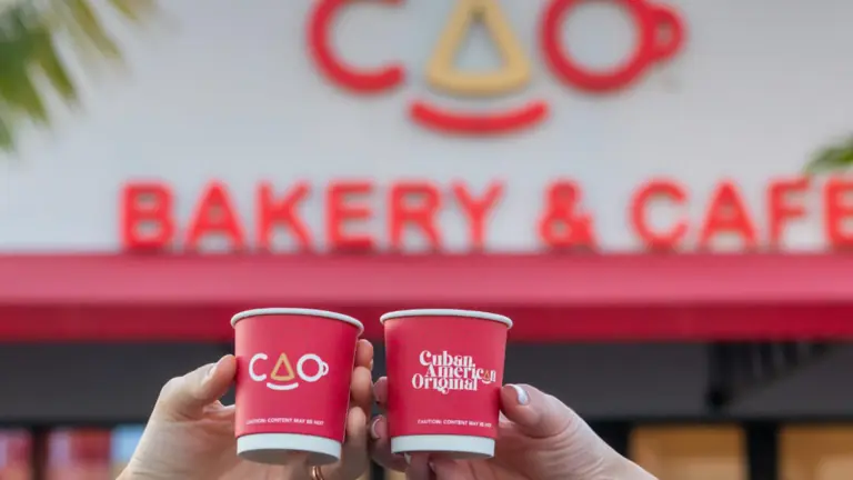 two cups of coffee held up in front of a large red bakery sign