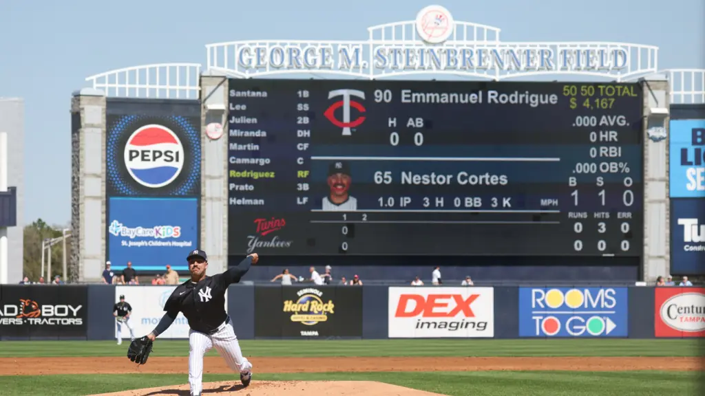 a pitcher at the mound throws the ball. A huge scoreboard is visible in the background