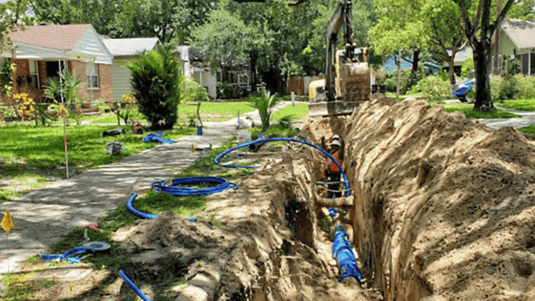 a large ditch dug out by trucks in a suburban neighborhood. Trees, and new pipes are being installed