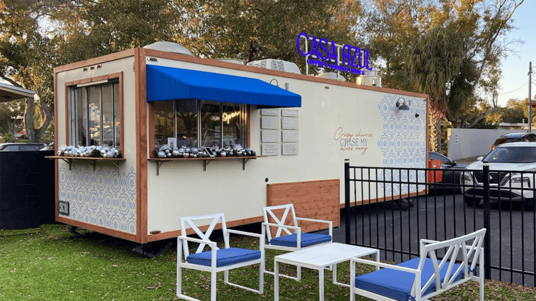 a food truck with a blue awning with turf surrounding the truck and tables arranged around it.