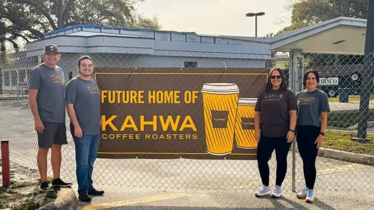 a group of people posed in front of a chain link fence with a brown and yellow sign announcing the arrival of a new coffee shop