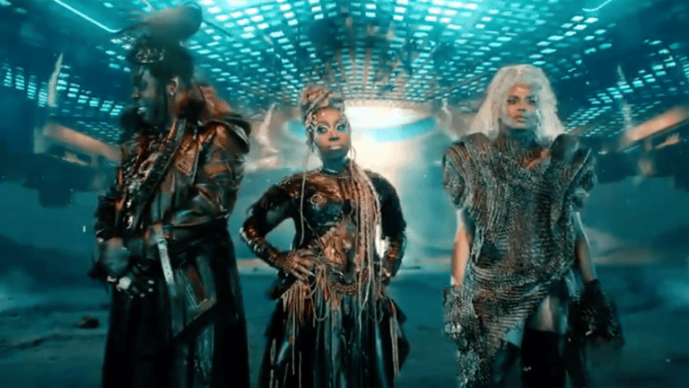 three people stand on a set that looks like outerspace. Each is adorned in gold body paint and futuristic outfits