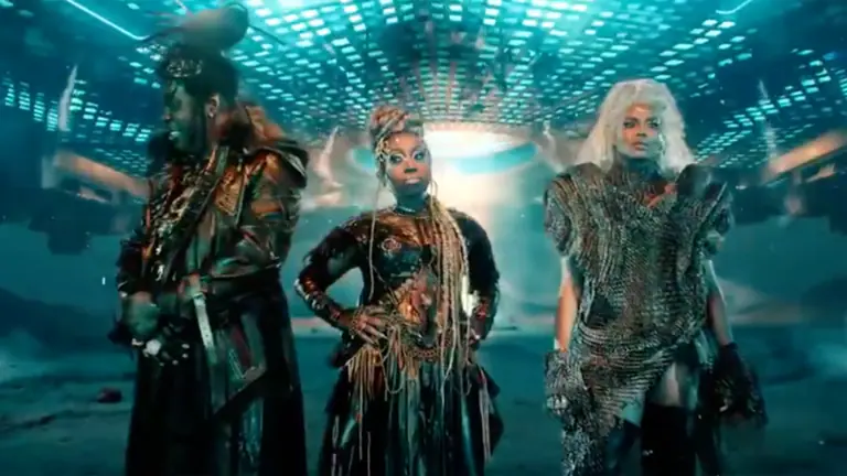 three people stand on a set that looks like outerspace. Each is adorned in gold body paint and futuristic outfits