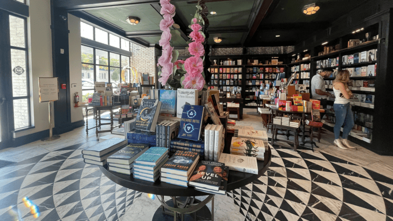 inside a bookstore with a checkerboard tile floor, and large plants placed on a center console with books stacked around them.