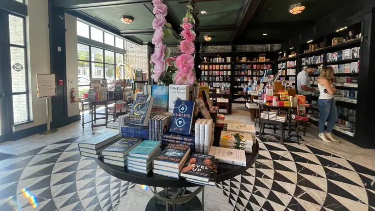 inside a bookstore with a checkerboard tile floor, and large plants placed on a center console with books stacked around them.