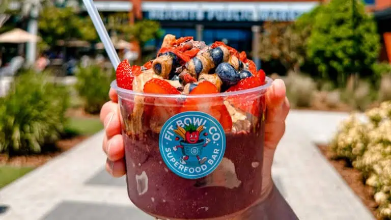 a smoothie bowl topped with blueberries and strawberries. A hand holds it up in front of a brick building on a sunny day