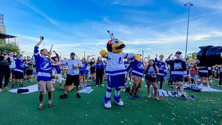 A group of people in blue shirts celebrate on a waterfront lawn with a thunder bug mascot