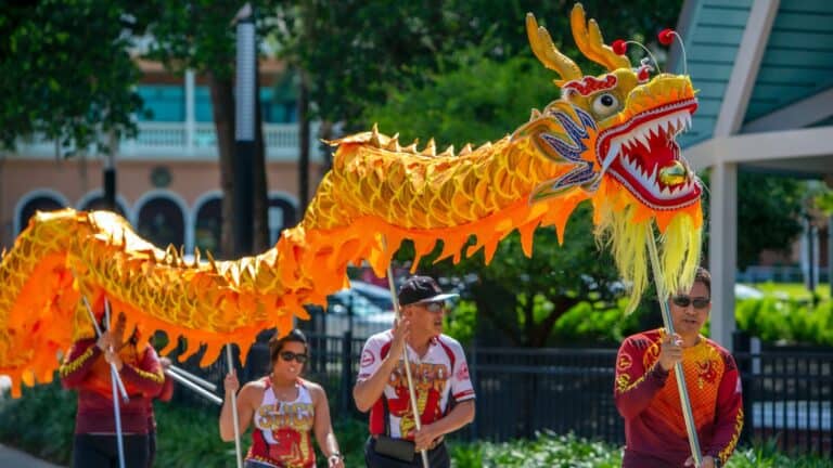 a group of people carry a paper dragon in a park. it's bright orange.