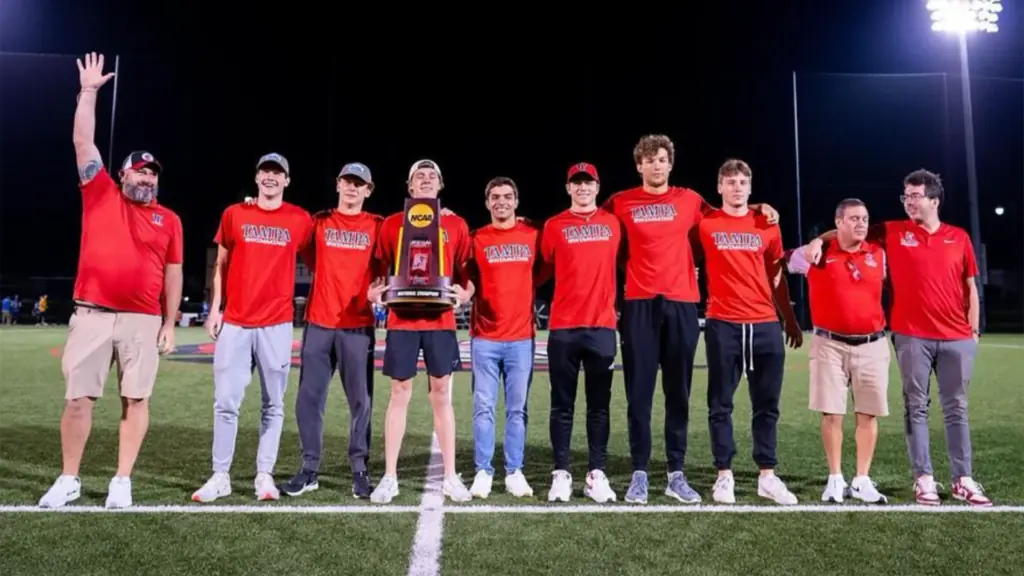 a group of athletes pose at the center of an athletic field at night - all wearing red shirts - and hold up a national championship trophy