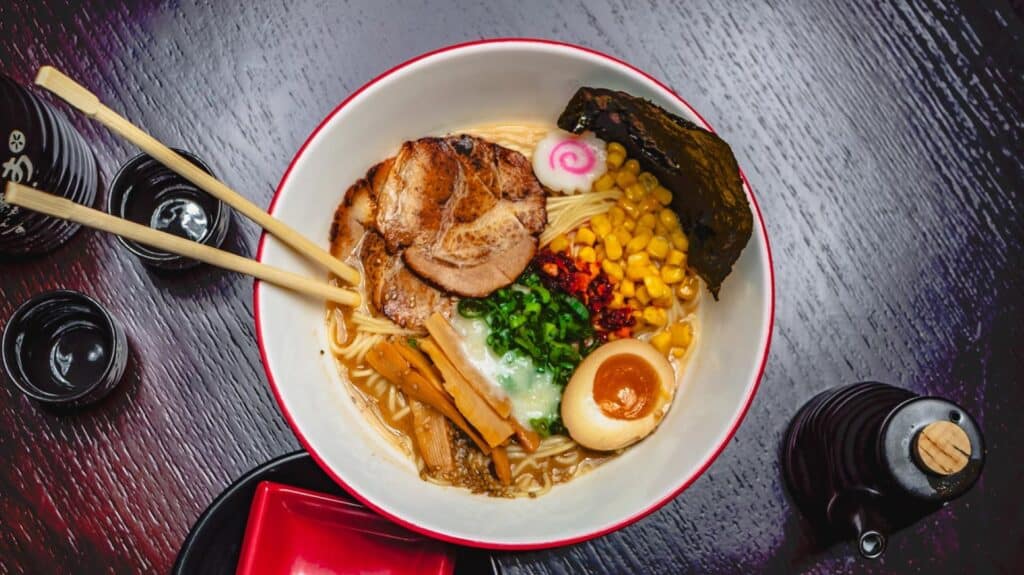 Photo of ramen bowl with chopsticks hanging on the side and a soy sauce bottle nearby