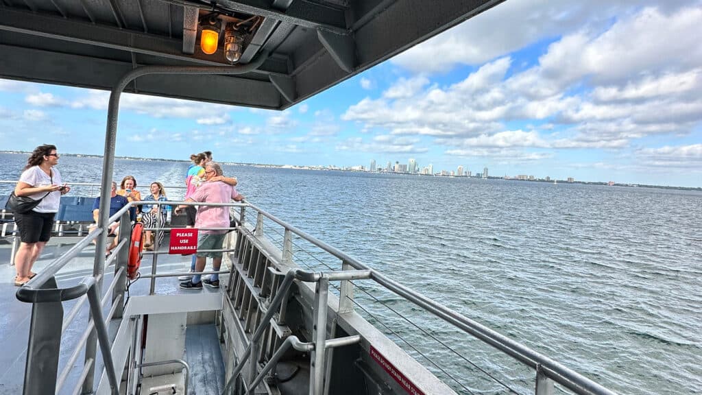 people stand on the upper deck of a ferry. A city skyline is visible on the horizon