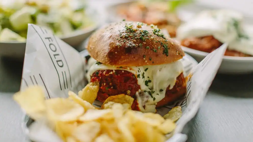 a small meatball sandwich covered in cheese. Chips are served on the side. The bun is covered in green garnishments